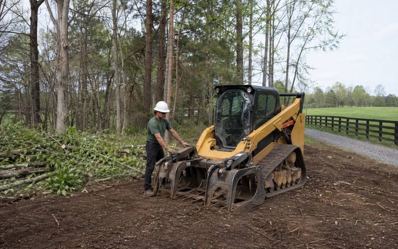 Worker guiding the loader's grounded grapple arm beside the windrow on a Madison tract part way through clearing.